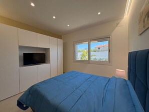 A bedroom with a bed, a TV mounted on the wall, a window with a view, and a lamp on the bedside table.