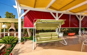 Ein überdachter Patio-Bereich mit einem gestreiften Gartenstuhl, einem Pizzaofen und Topfpflanzen.