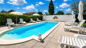 Una piscina con sillas de descanso y sombrillas, rodeada por un seto y una pared blanca.