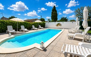 Ein Schwimmbecken mit Liegestühlen und Sonnenschirmen, umgeben von einer Hecke und einer weißen Mauer.