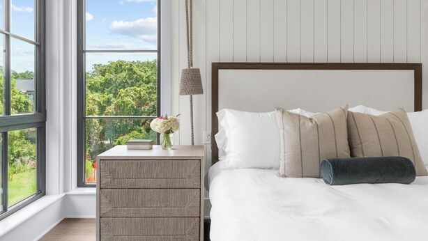 Une chambre avec une grande fenêtre, un lit recouvert de draps blancs et agrémenté d’oreillers à rayures, une table de chevet avec une lampe, et une vue sur la verdure à l’extérieur.