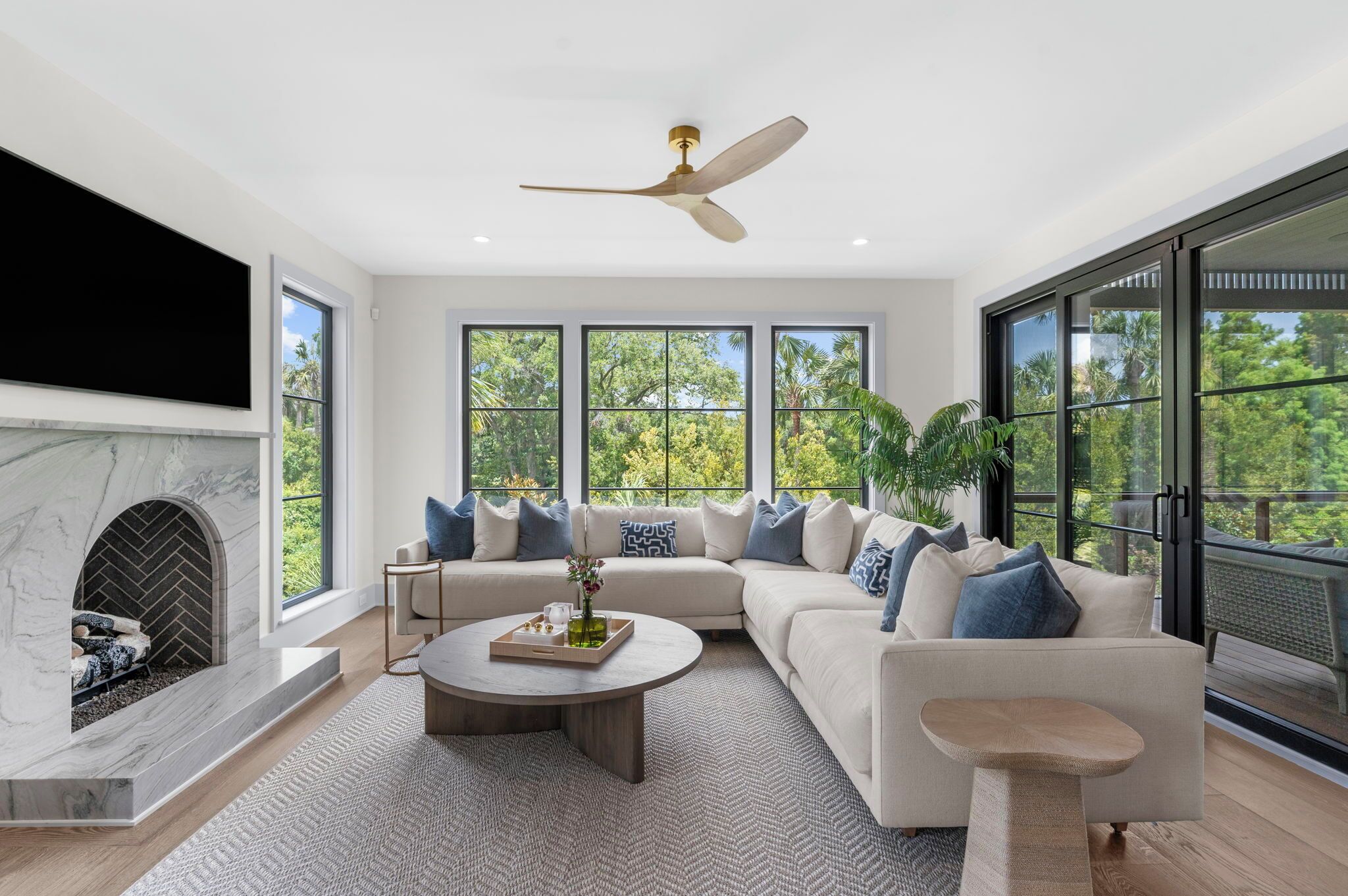 Un salon moderne doté d’un canapé d’angle, d’une cheminée, d’un ventilateur de plafond et de grandes fenêtres offrant une vue sur la verdure.