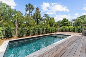 Ein rechteckiger Swimmingpool mit klarem Wasser, umgeben von einer Holzterrasse und vielfältigem Grün, darunter Palmen und Sträucher.