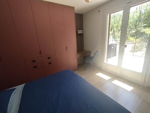Ein Schlafzimmer mit einem Bett, einem Schreibtisch und einem Stuhl. Eine Glastür führt zu einem Außenbereich.