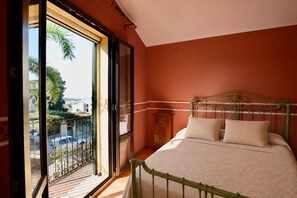 Ein Schlafzimmer mit großem Fenster, einem Bett mit Kissen, einem Nachttisch und einem Holzkasten.