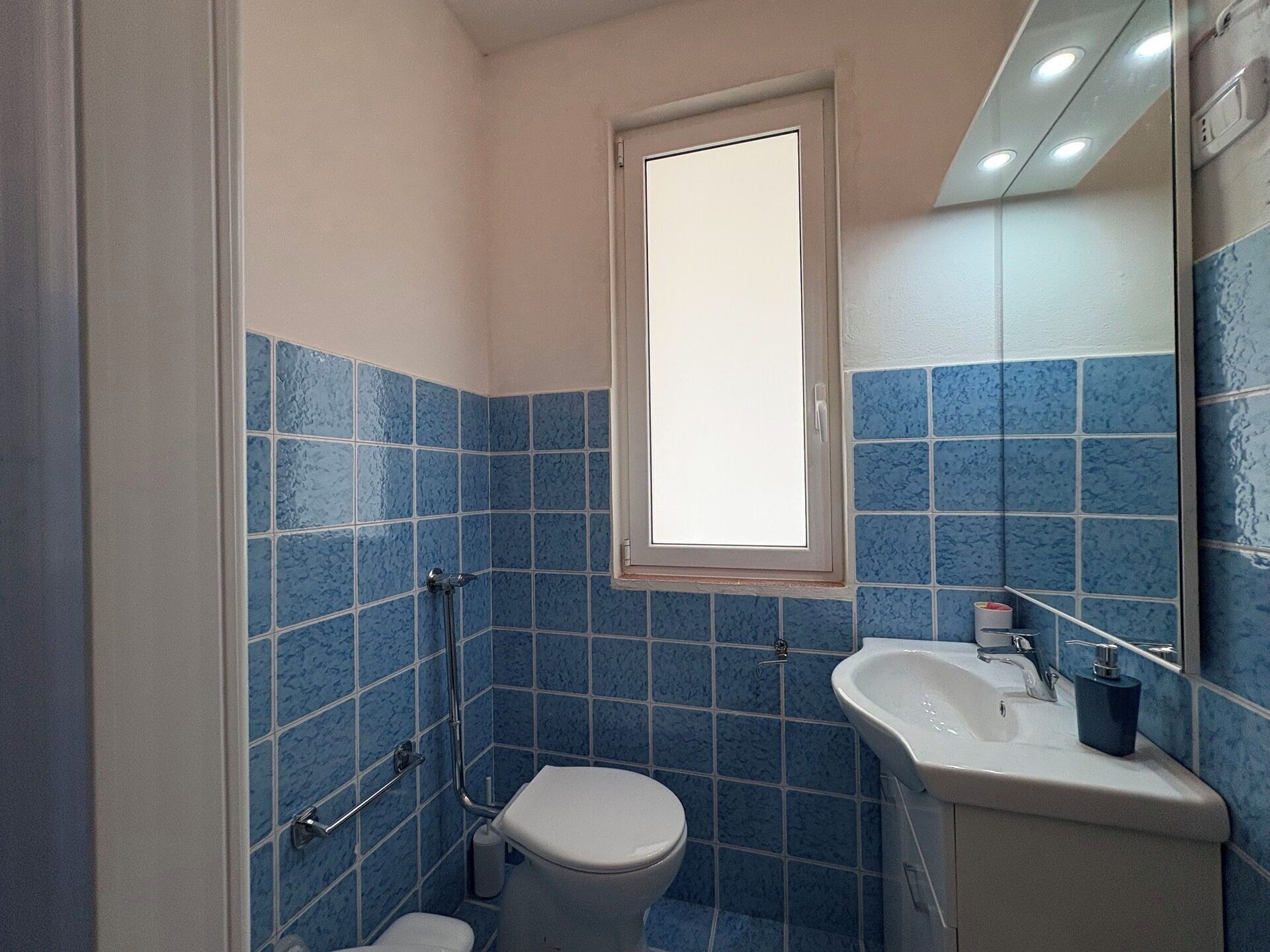 Ein Badezimmer mit blauen Fliesen, einer weißen Toilette, einem Waschbecken und einem Spiegel.