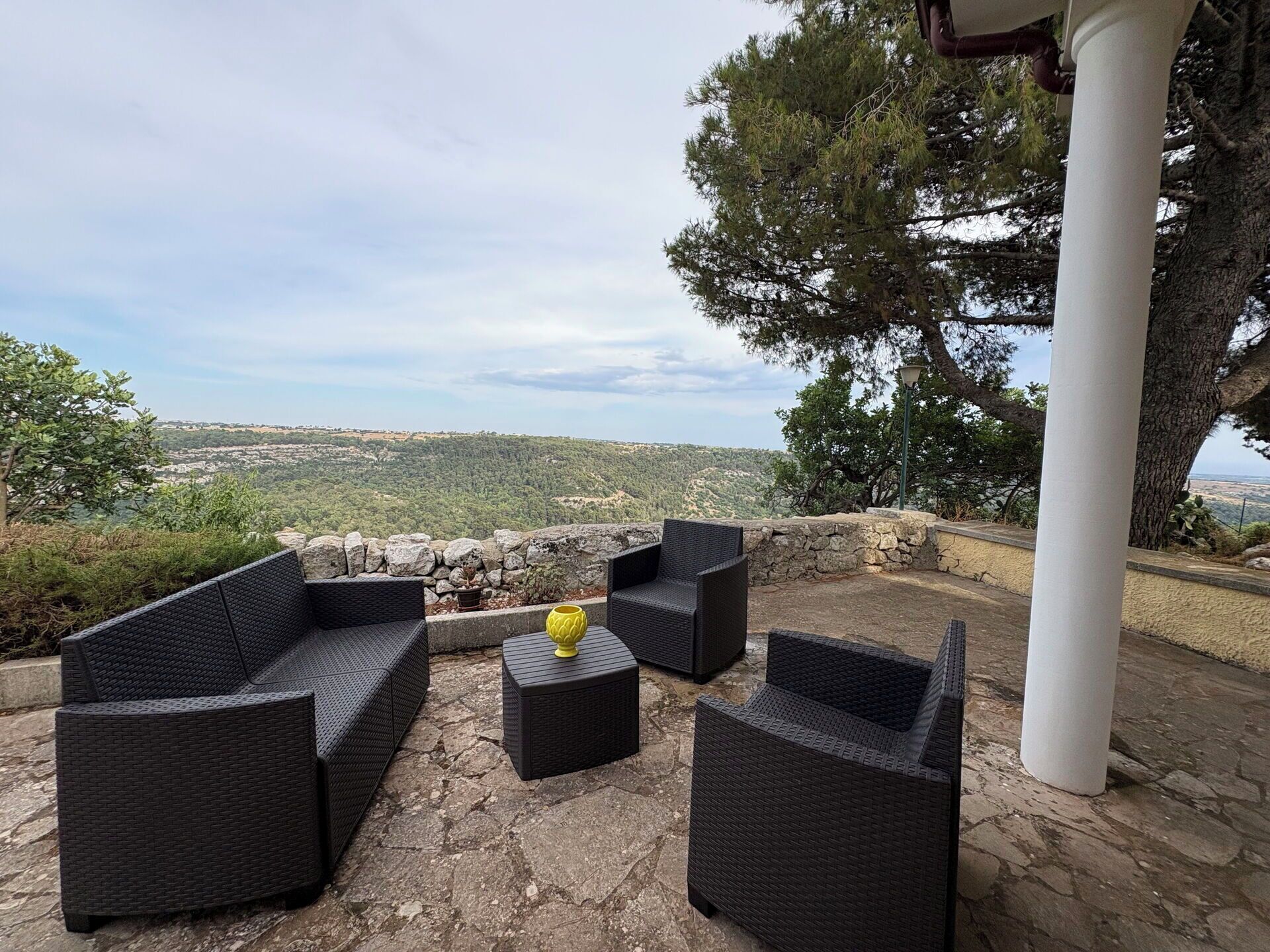 An outdoor seating area with wicker furniture and a stone wall, overlooking a hilly landscape.