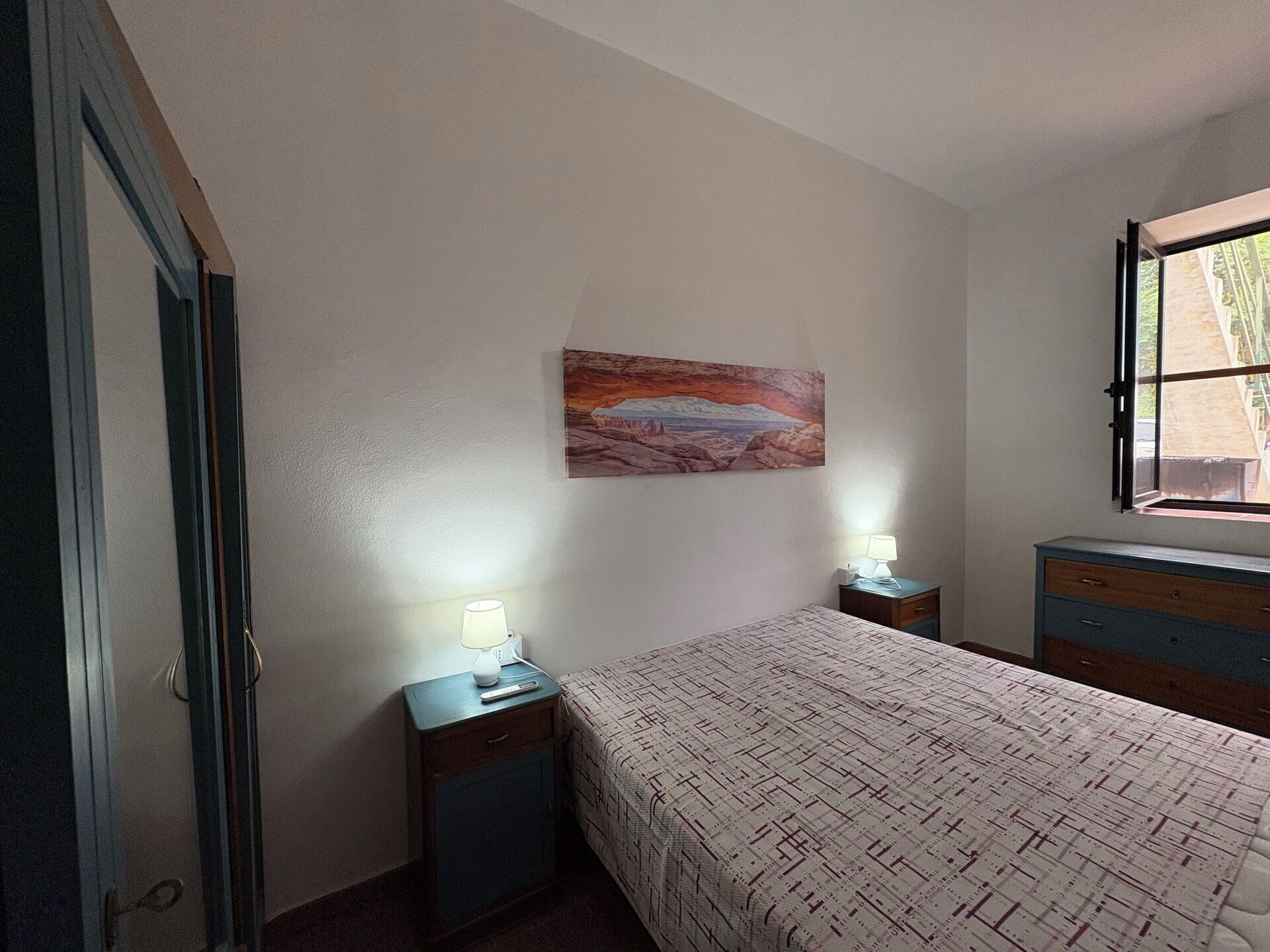 A bedroom with a bed, a nightstand, a dresser, a painting on the wall, and a window.