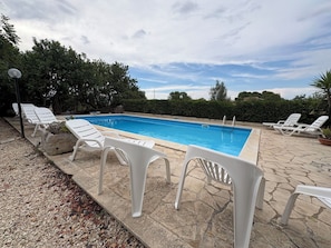 Ein Pool mit weißen Liegestühlen, umgeben von Steinplatten und Grünpflanzen.
