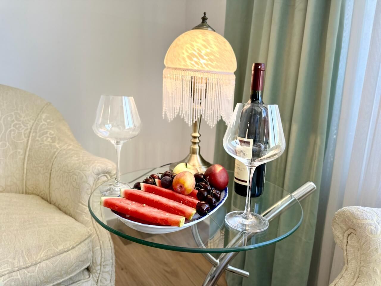 Una mesa de vidrio con una botella de vino, dos copas y un plato de fruta.