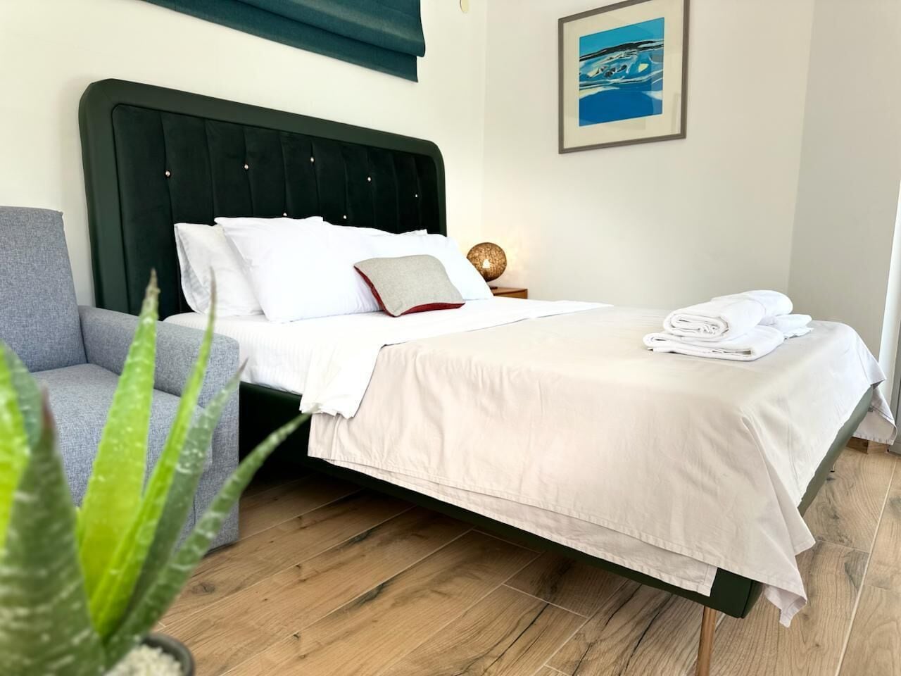 Ein ordentlich bezogenes Bett mit weißen Leinen, ein graues Sofa, Holzboden und ein gerahmtes Bild an der Wand.