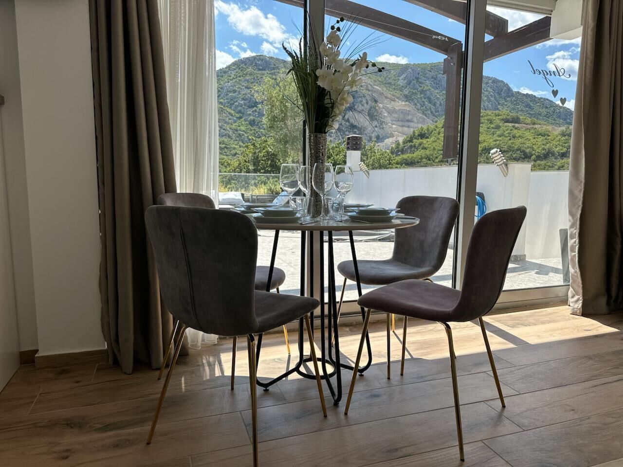 A dining area with a table and four chairs, a vase with flowers, and a view of mountains through large windows.