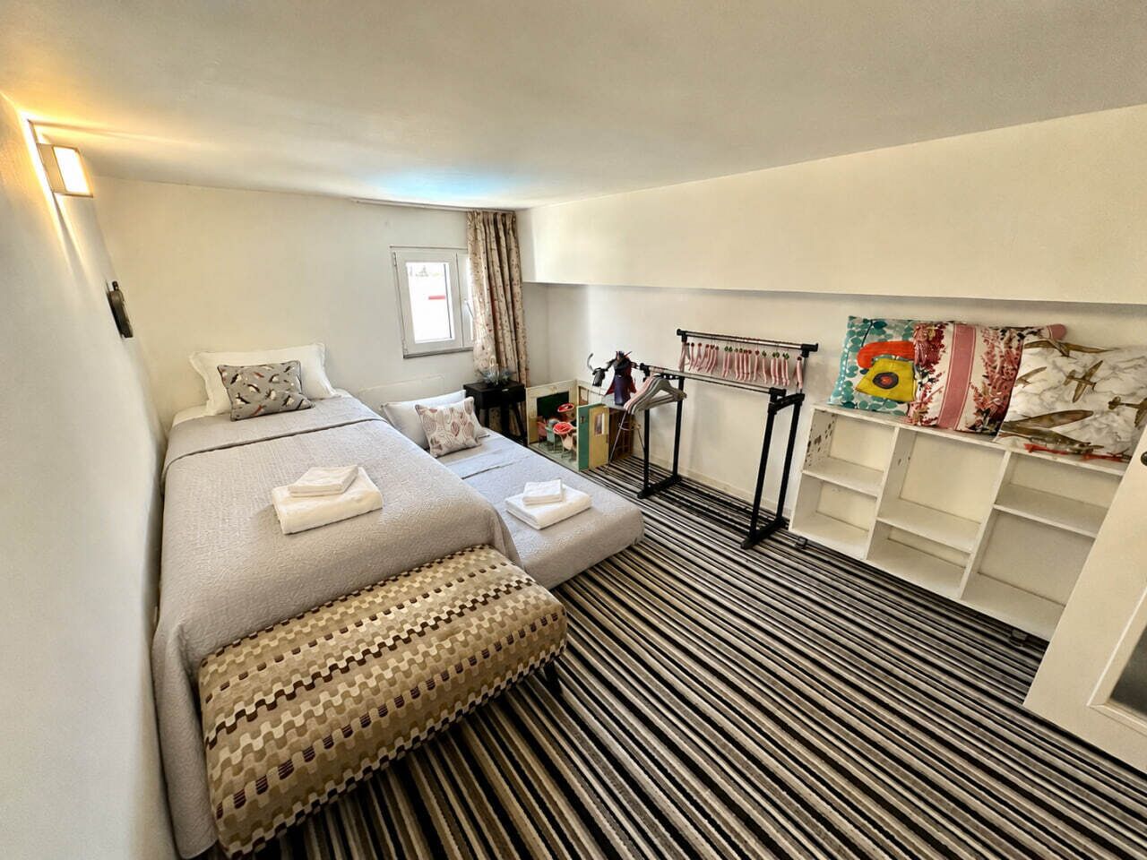 A hotel room with a bed, a bedside table, a lamp, a window with curtains, and a storage unit with shelves.