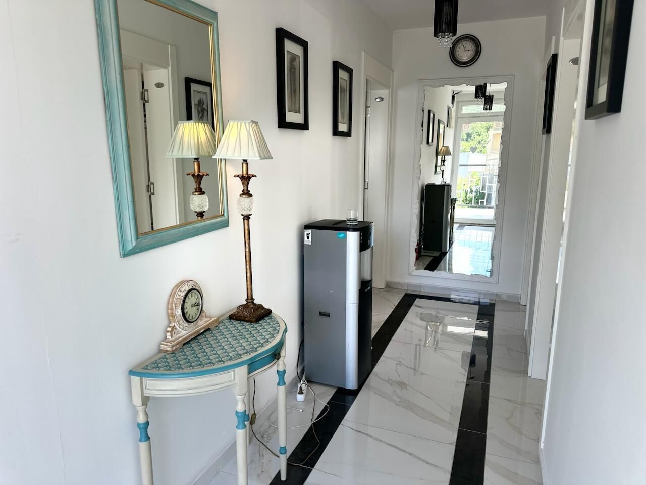 A hallway with a mirror, two wall-mounted lamps, a small table with a clock, and a refrigerator.