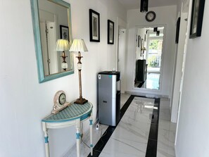 Un pasillo con un espejo, dos lámparas de pared, una mesita con un reloj y un refrigerador.