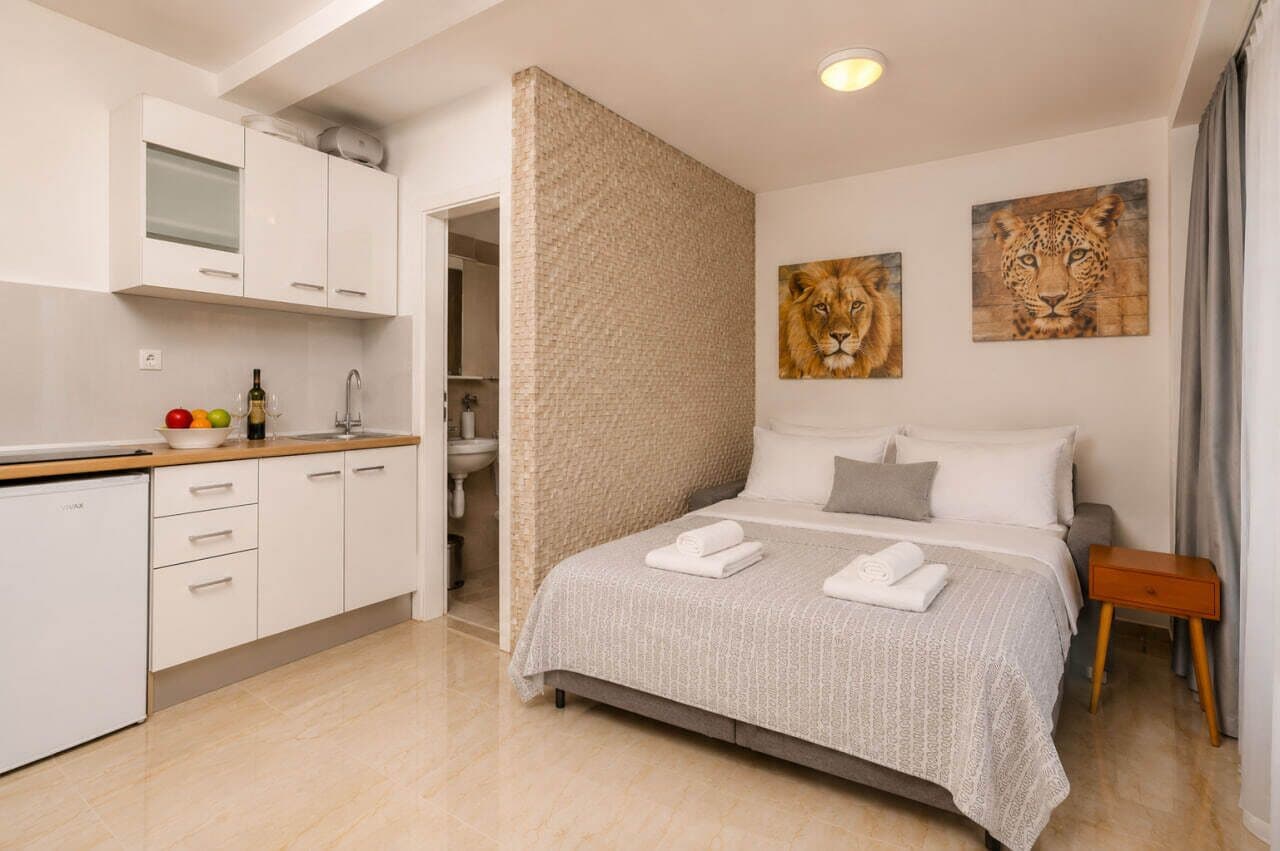 A modern hotel room with a bed, a small bedside table, a wall-mounted painting of a lion, and a kitchen area with cabinets and a countertop.