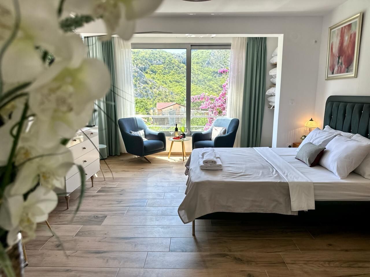 A hotel room with a bed, two chairs, a small table with a bottle, and a view of greenery through large windows.