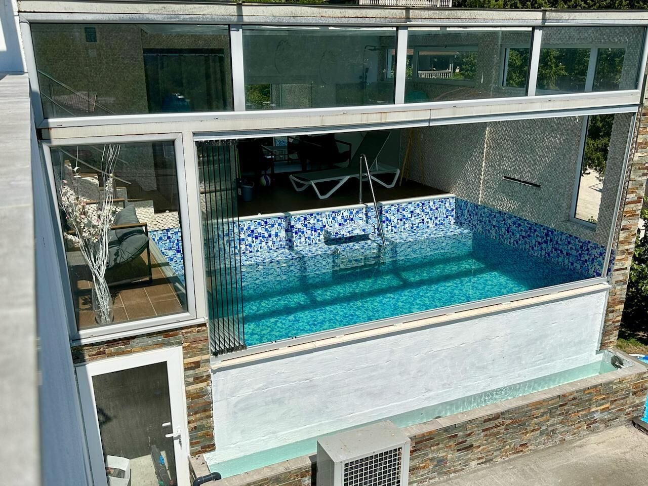 Ein Schwimmbecken mit einer gefliesten Kante, einer Glaskonstruktion und einer Steinmauer.