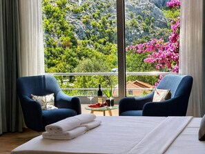 A hotel room with a bed, two armchairs, a small table with a bottle and glasses, and a balcony with a view of greenery and flowers.