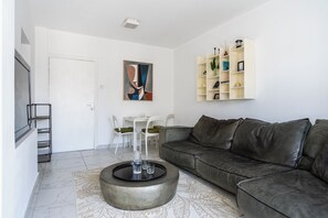 Un saló modern amb un sofà de cuir gris, una taula de centre rodona, una porta blanca, una paret blanca amb un quadre abstracte emmarcat i un moble prestatge blanc amb objectes decoratius.