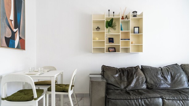 Ett modernt vardagsrum med ett vitt matbord, två vita stolar, en grå lädersoffa och en vägghängd hylla med olika dekorativa föremål.