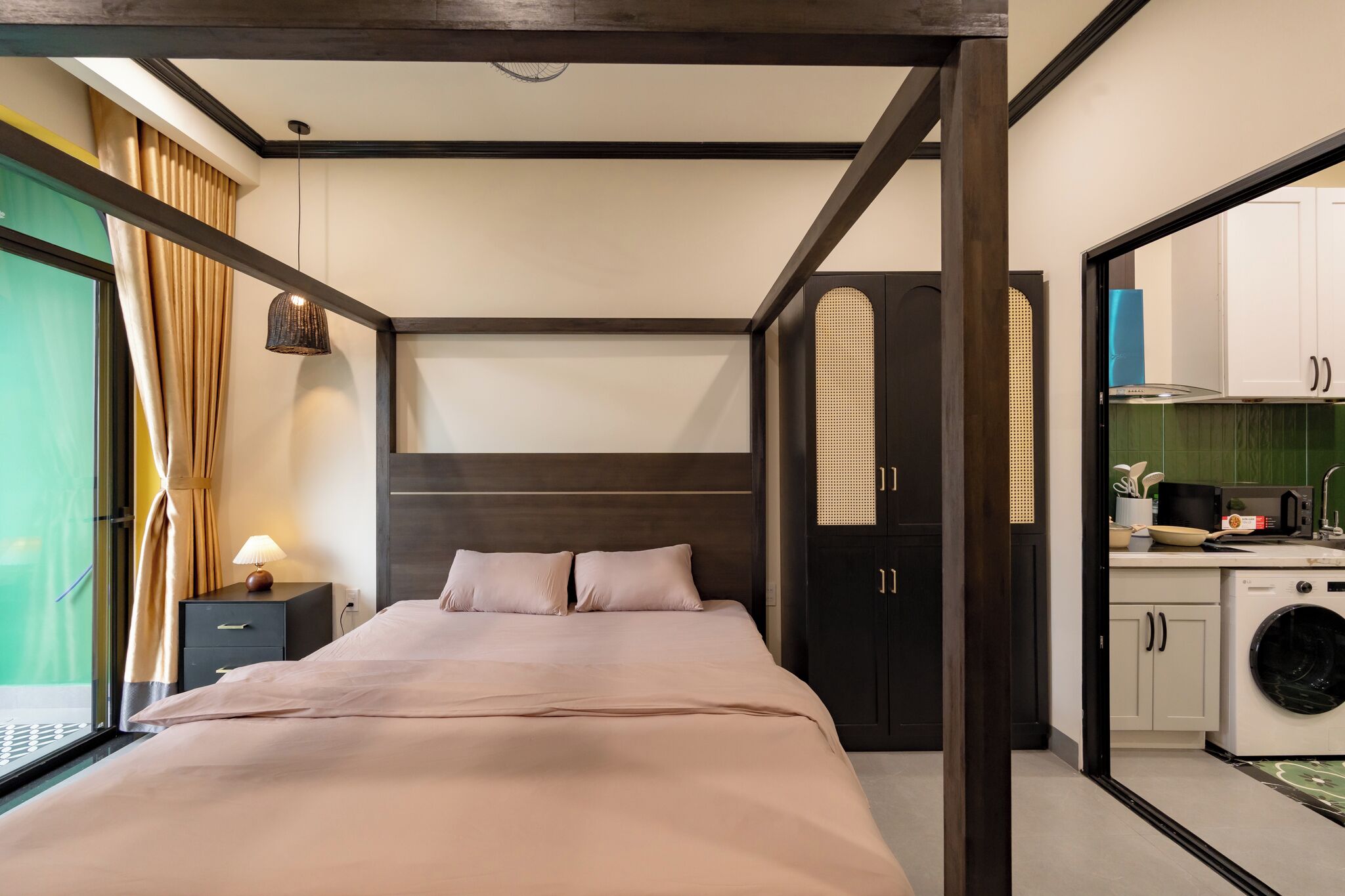 A bedroom with a bed, bedside table, lamp, and a view of a kitchen area with a washing machine.