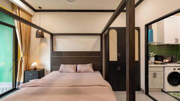 A bedroom with a bed, bedside table, lamp, and a view of a kitchen area with a washing machine.