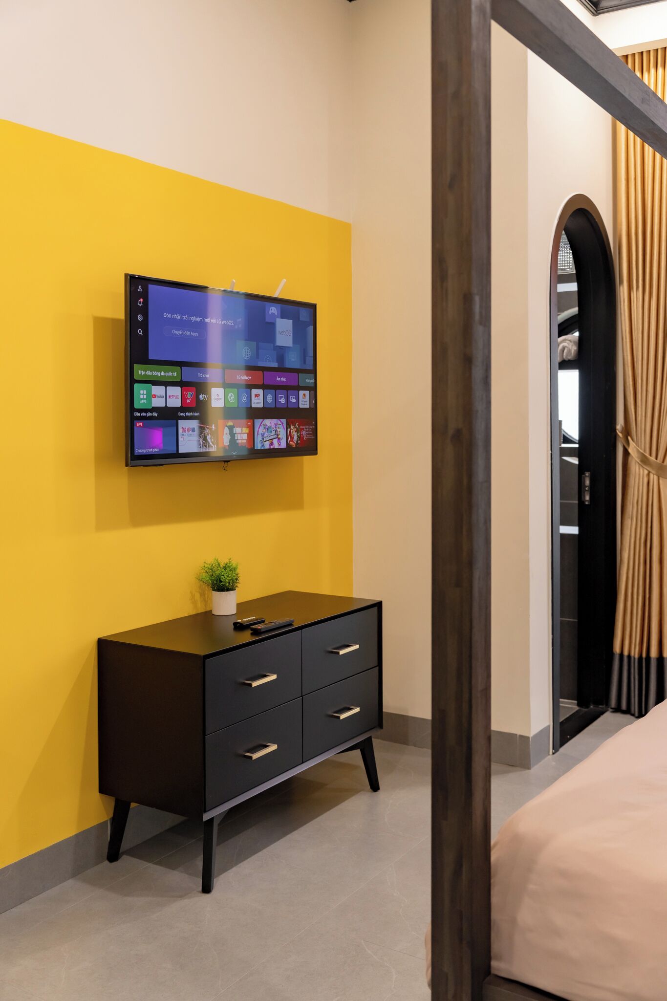 Quarto moderno com televisão na parede, cómoda preta com gavetas e cama com cobertura bege.