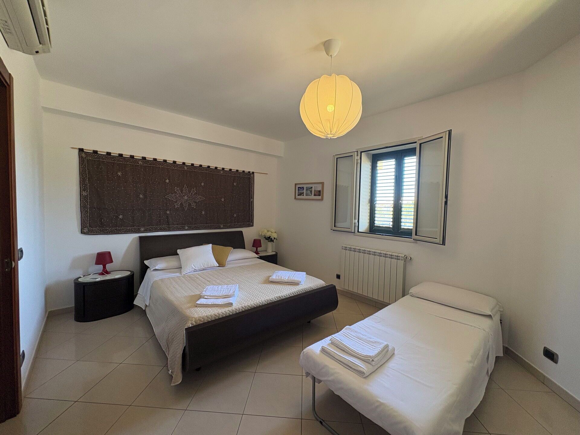 A bedroom with a large bed, a smaller bed, a nightstand, a lamp, a radiator, a window with blinds, and a hanging light fixture.