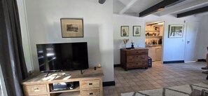 A living room with a flat-screen TV on a wooden stand, a wooden cabinet, and a kitchen area with a refrigerator and cabinets. - OASE AM DEICH (Krummendeich)