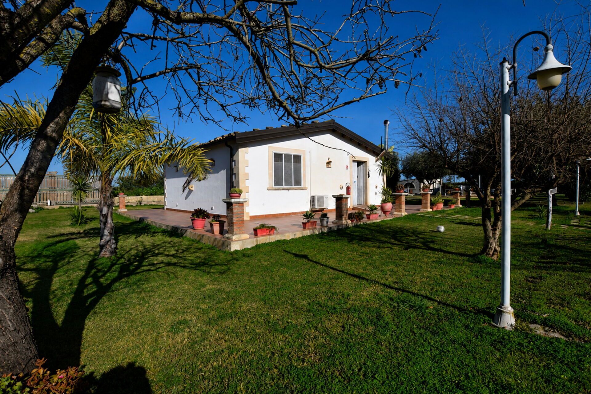 A single-story house with a white exterior, a small porch with potted plants, and a clear blue sky.