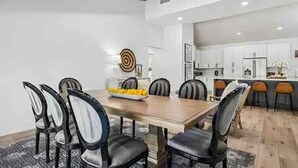 A modern dining area with a wooden table and black chairs, a kitchen in the background, and a decorative wall piece. - The Rally House-Premium 4 bed, priv pool, pickleball, 5 hole golf — mins to TPC (Scottsdale)