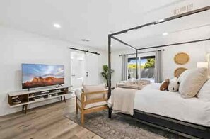 A modern bedroom with a four-poster bed, a television on a stand, and a view of the outdoors through a window. - The Rally House-Premium 4 bed, priv pool, pickleball, 5 hole golf — mins to TPC (Scottsdale)