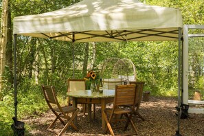 Un espace de restauration en plein air, avec une table et des chaises placées sous une tonnelle, le tout entouré d’arbres.