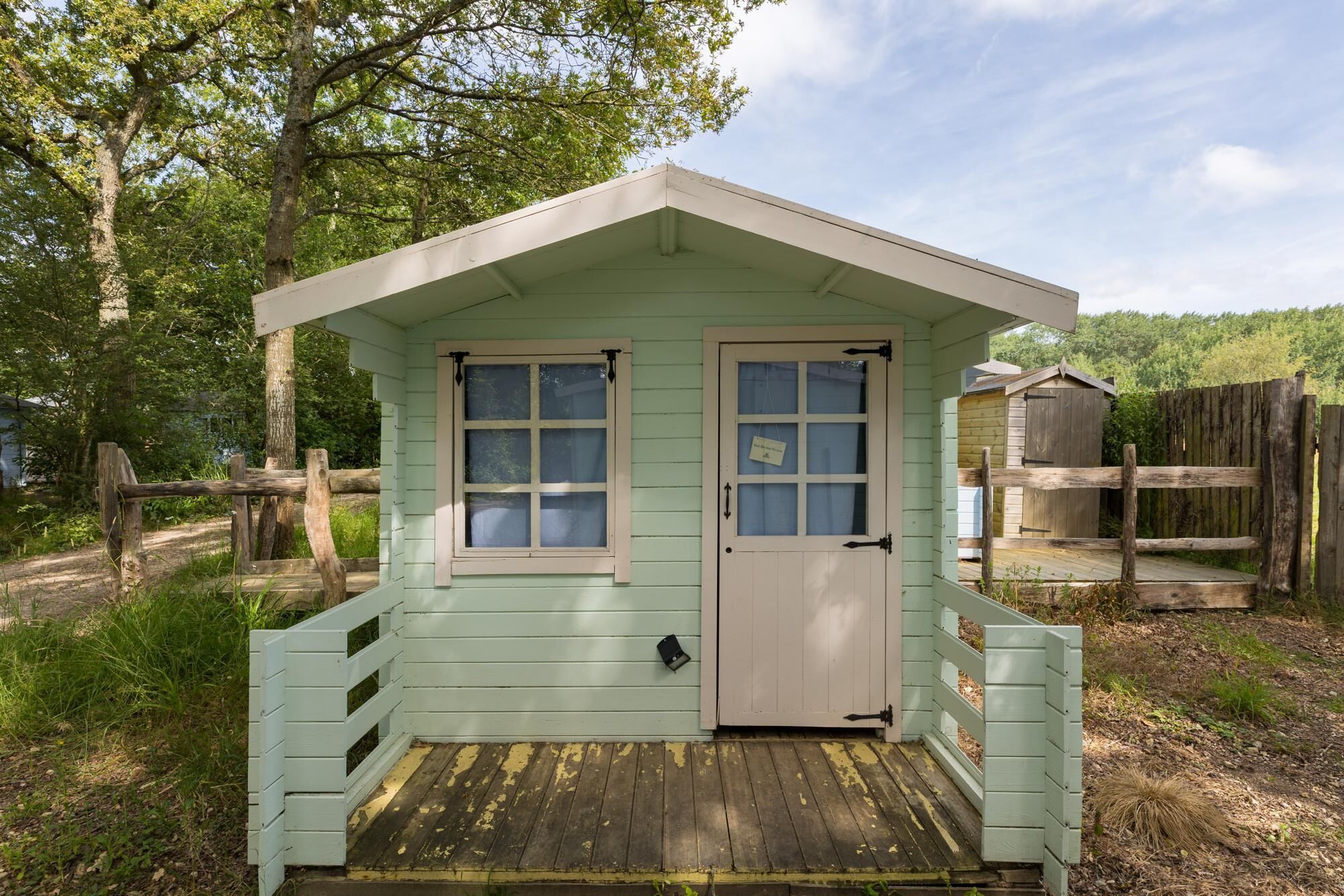 A small, light green wooden structure with a porch and two windows, surrounded by trees and a wooden fence.