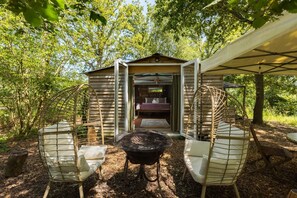 A rustic wooden cabin with a bed, a grill, and two wicker chairs. - Stunning Glamping Retreat in the Woods - Pass the Keys (Moreton-in-Marsh)