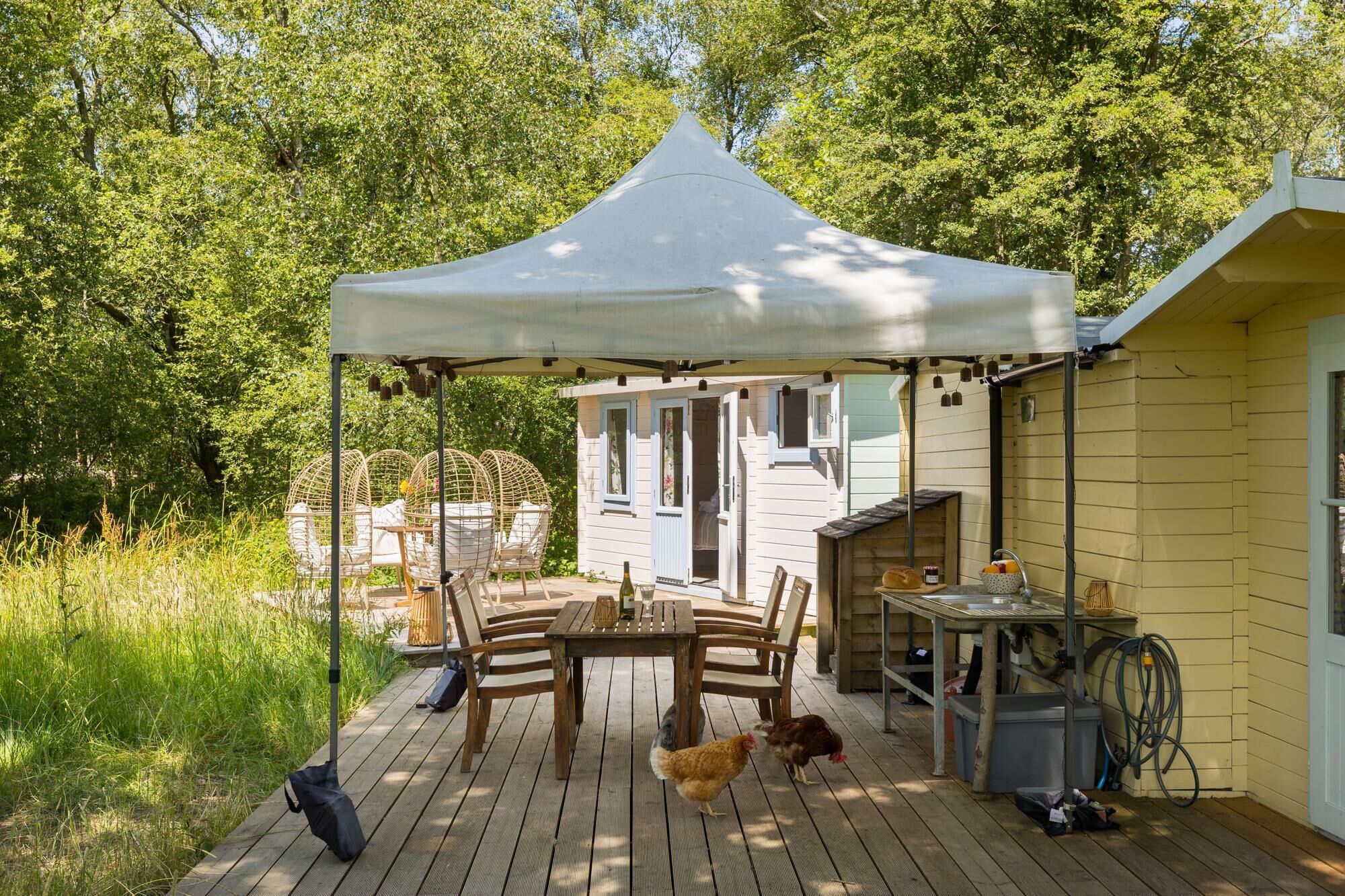 Een houten terras met een tafel, stoelen, een witte tent en een klein gebouw met een deur en ramen.