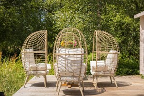 Three wicker chairs with cushions, arranged on a wooden deck with a garden backdrop.