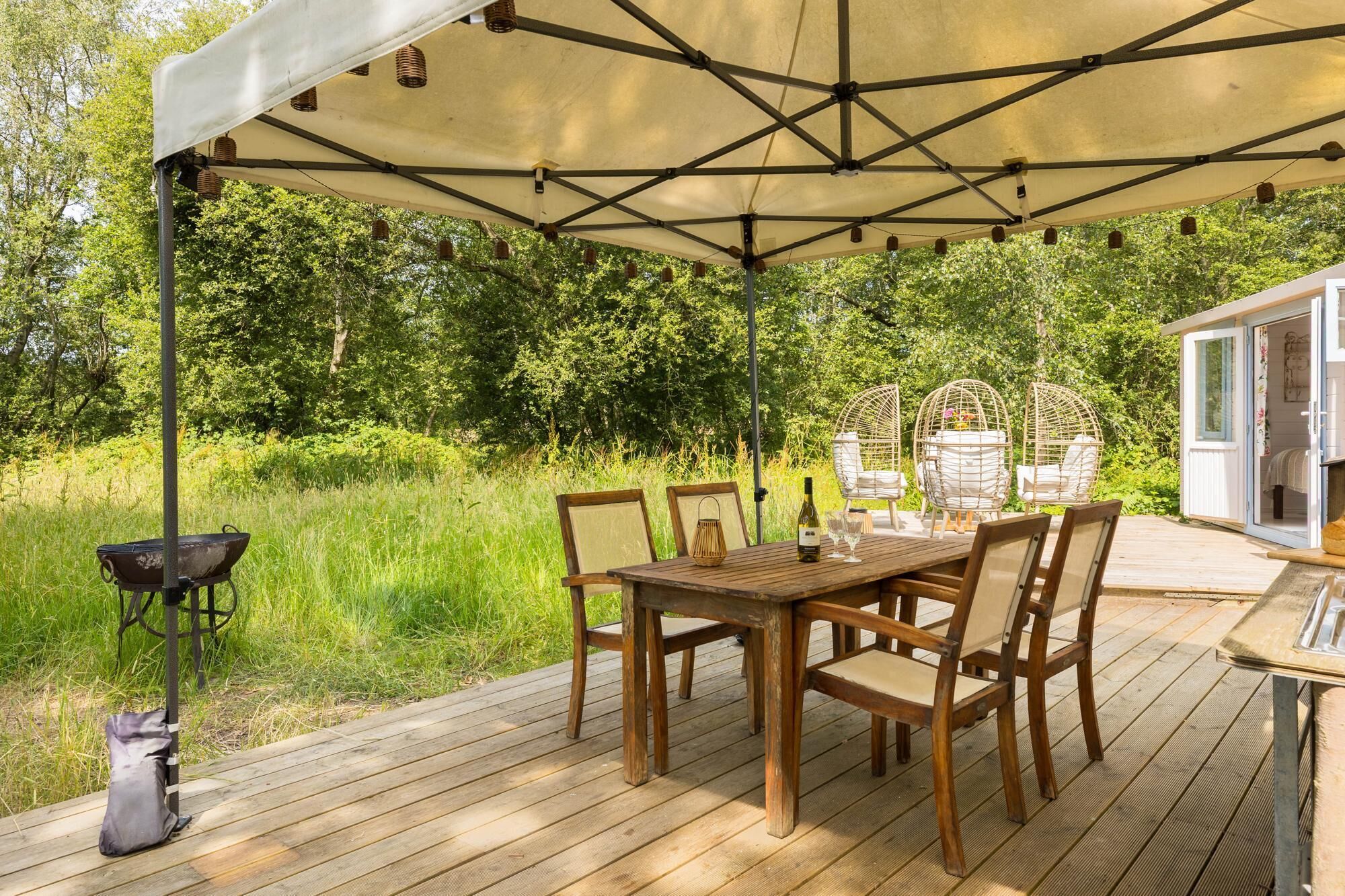 Une terrasse extérieure en bois, avec une table et des chaises placées sous une tonnelle, le tout entouré de verdure.