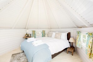 Um quarto estilo tenda, aconchegante, com teto branco, cama, mesas de cabeceira e uma luminária.