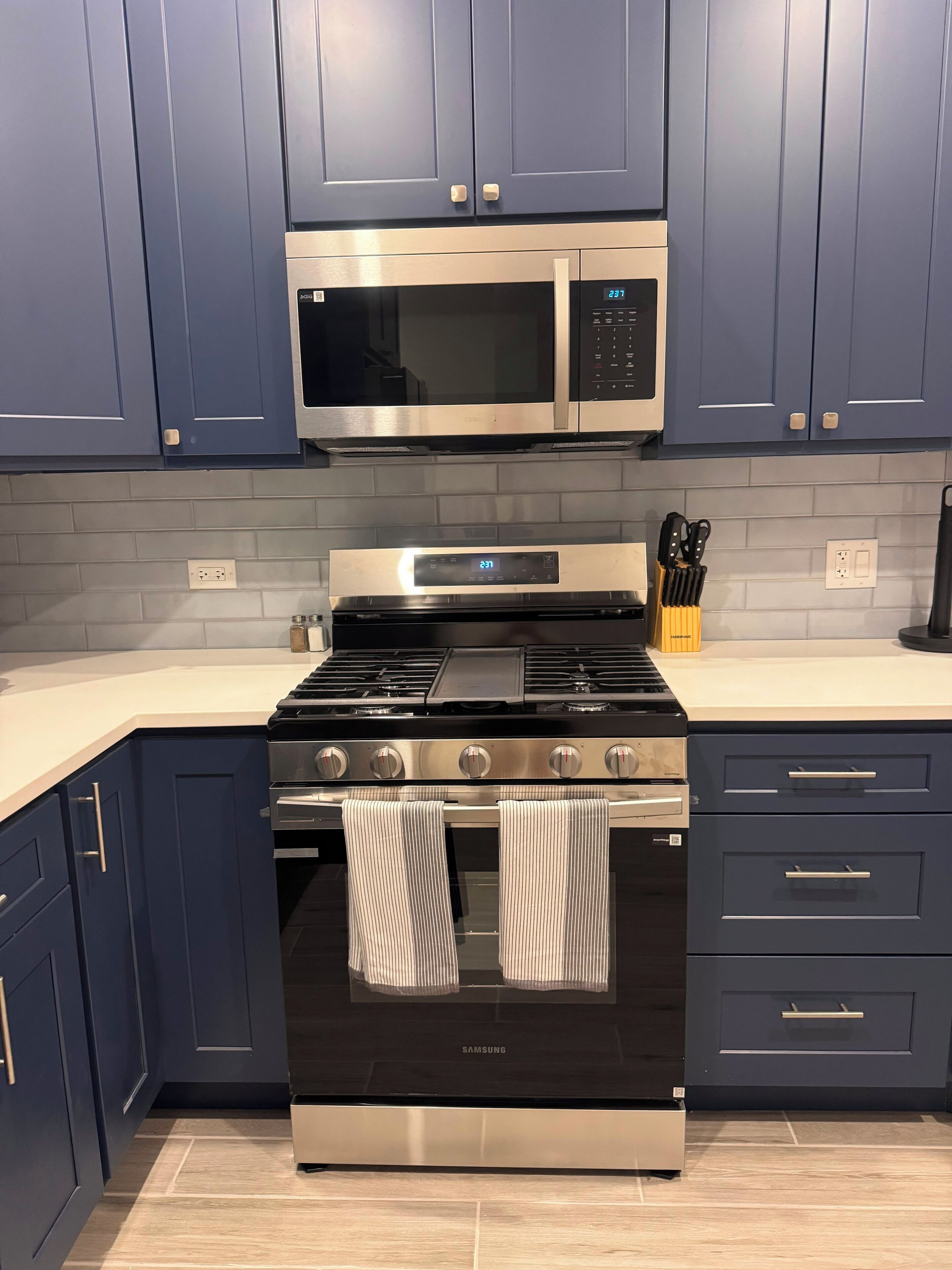 A kitchen with dark blue cabinets, a stainless steel microwave, a black Samsung oven, and a countertop with kitchen utensils.