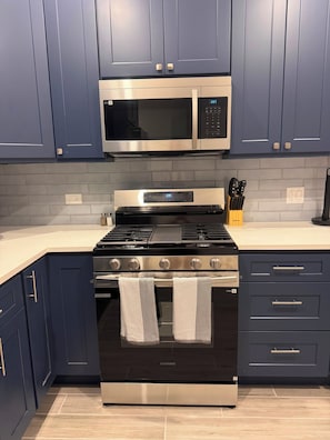 A kitchen with dark blue cabinets, a stainless steel microwave, a black Samsung oven, and a countertop with kitchen utensils.