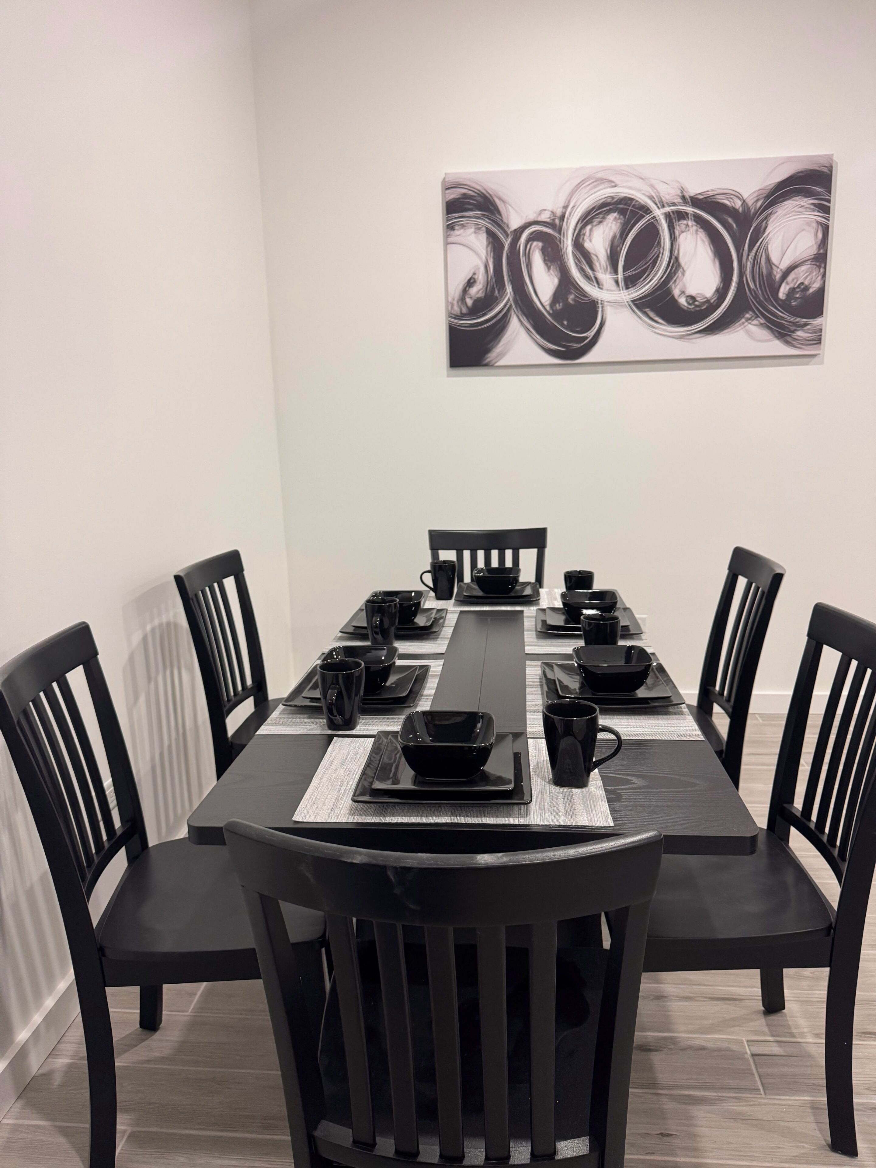 Ein Essbereich mit einem langen Tisch, gedeckt für vier Personen, umgeben von schwarzen Stühlen, mit einem modernen, abstrakten Gemälde an der Wand.