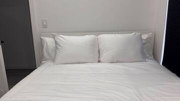 Uma cama com roupa de cama e almofadas brancas, uma parede lisa e uma porta à esquerda.