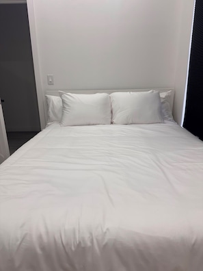A bed with white bedding and pillows, a plain wall, and a door to the left.