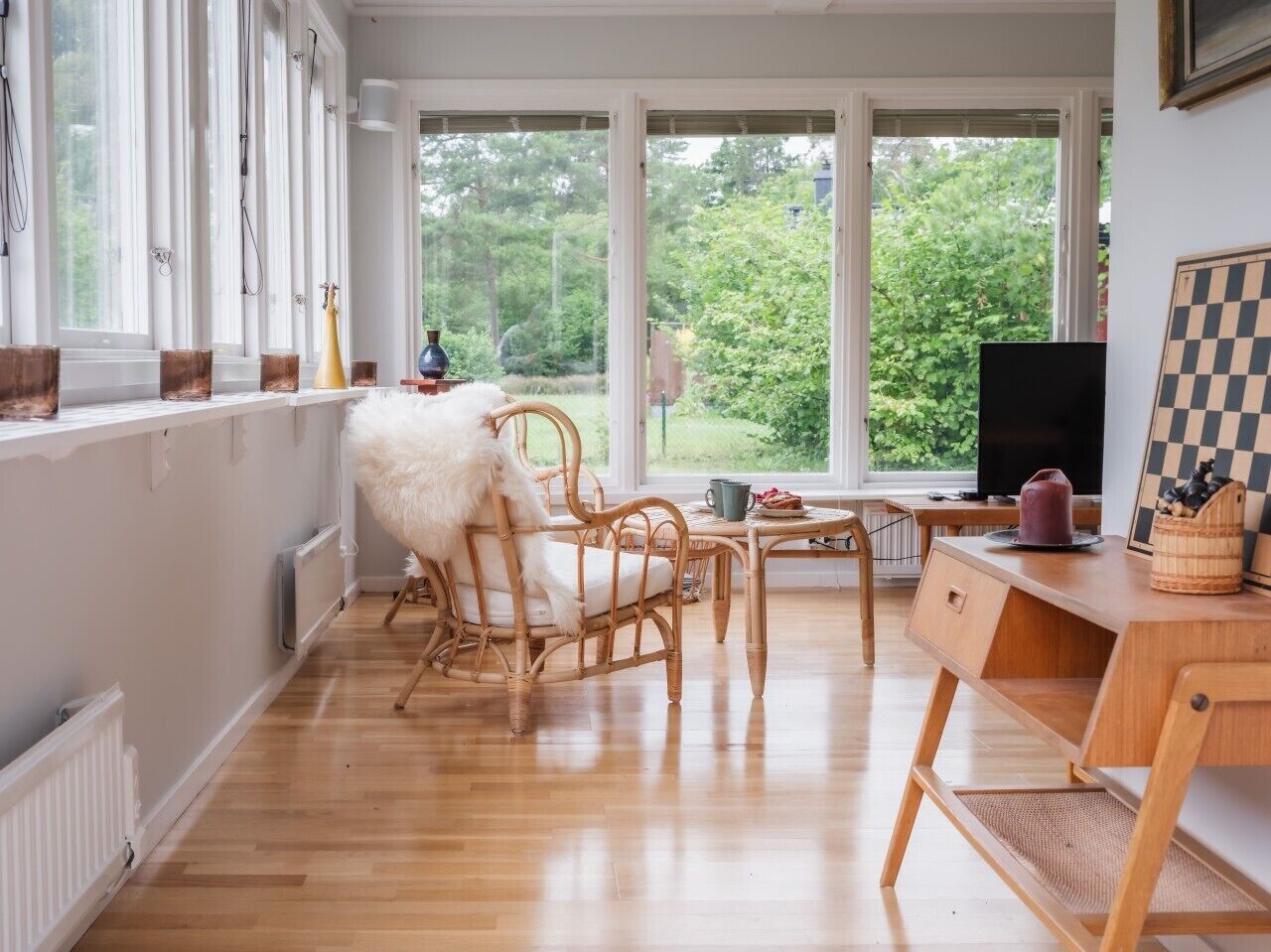 A room with a wooden floor, a white fluffy chair, a wooden desk, and a window with a view of greenery.