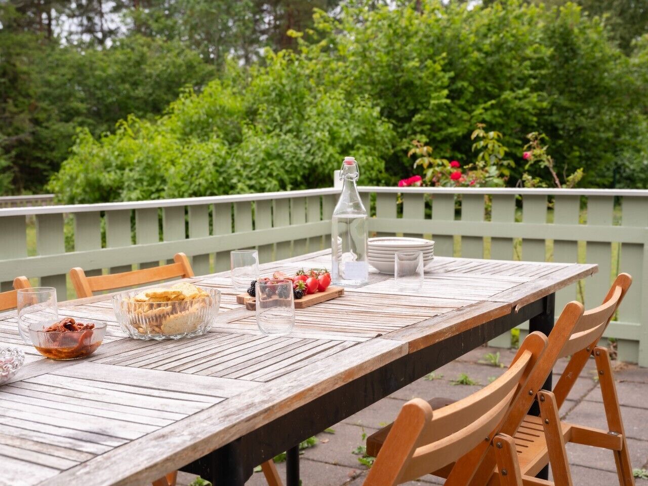 A wooden outdoor dining table with chairs, surrounded by greenery and a white fence.