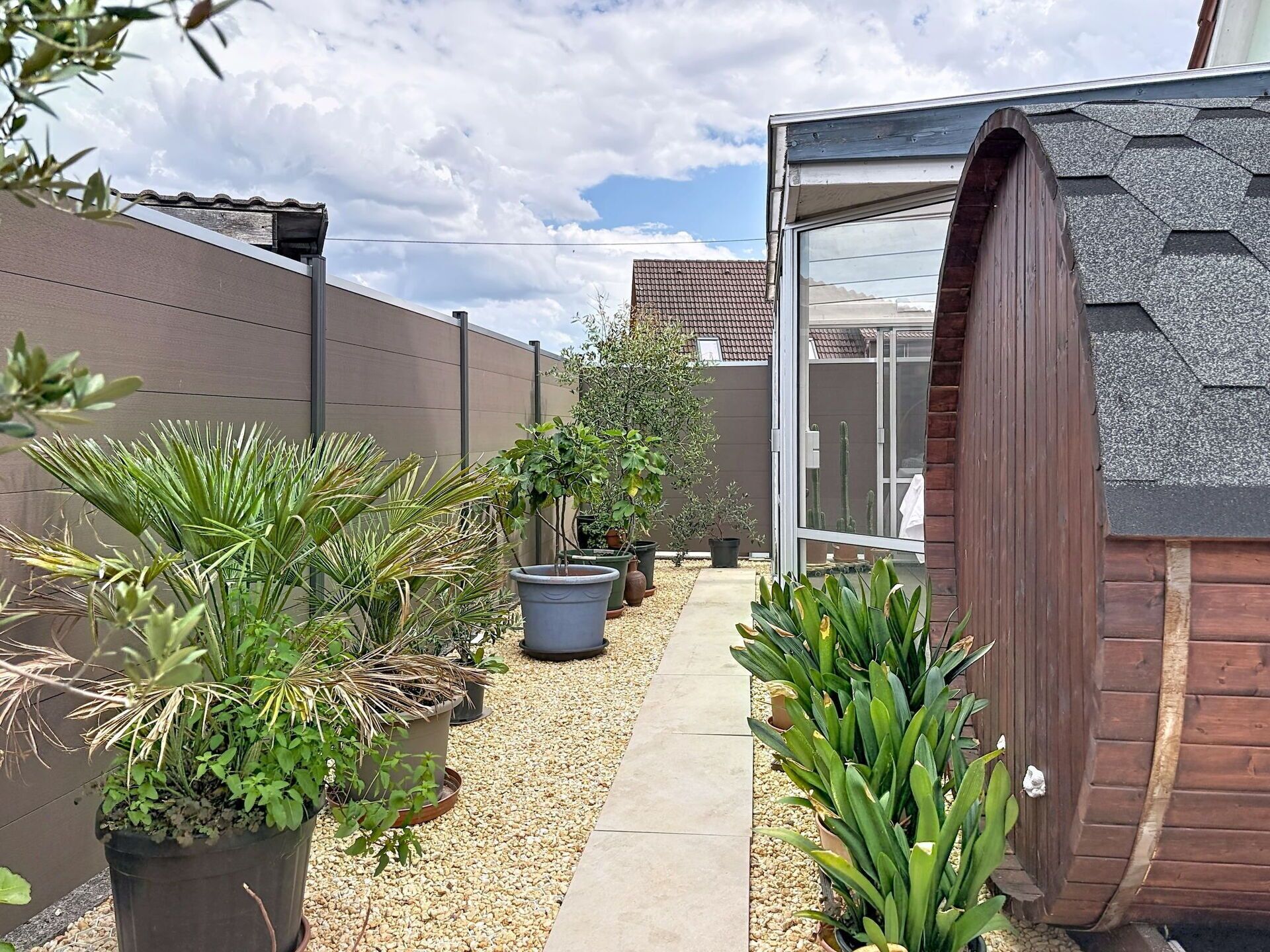 Un étroit sentier bordé de plantes en pot et d’une clôture en bois, qui mène à un bâtiment doté d’un toit en pente.
