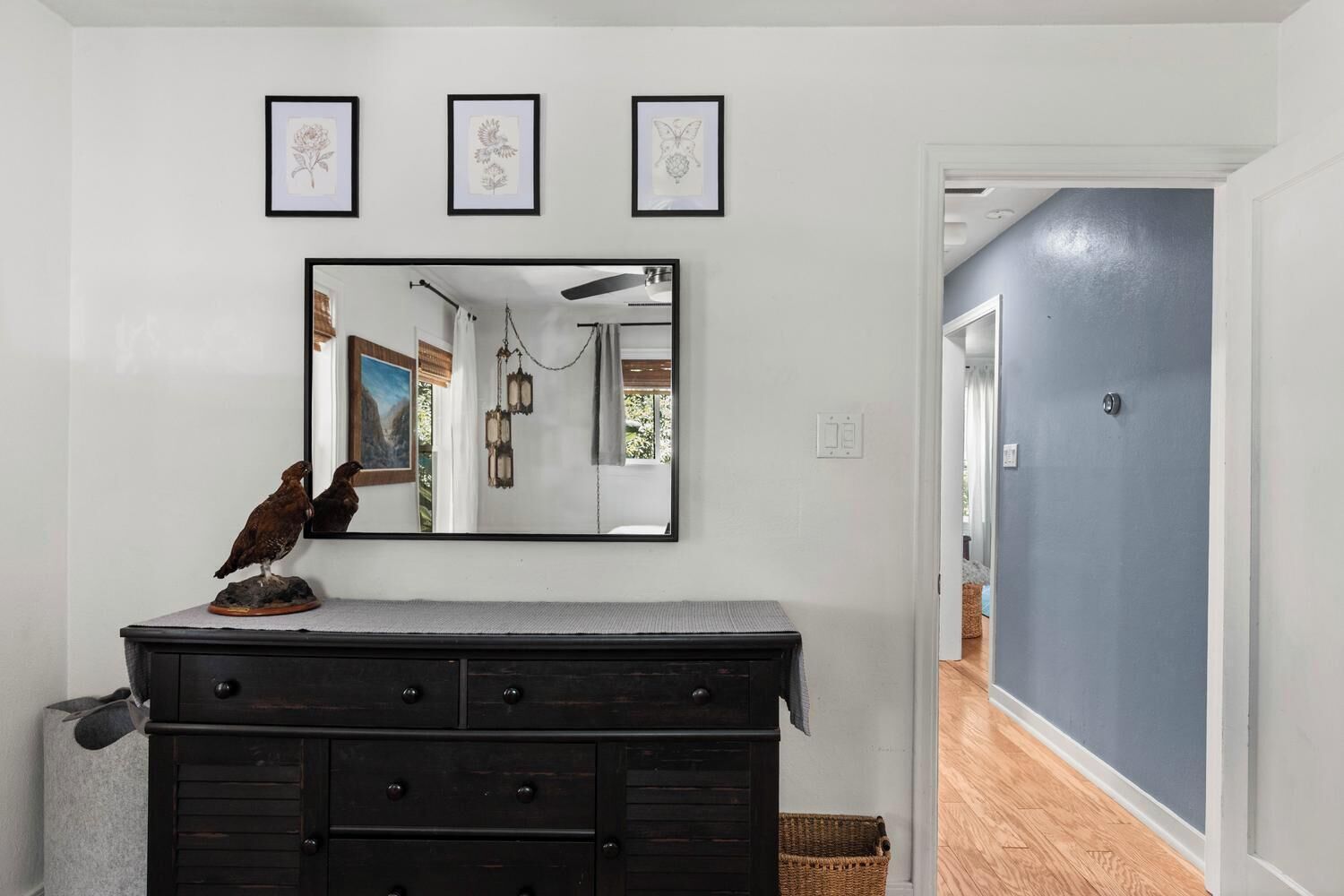 A room with a dark wooden dresser, a wall-mounted mirror reflecting a hallway, and three framed botanical prints above the mirror.