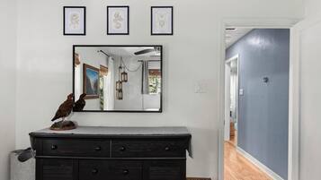 A room with a dark wooden dresser, a wall-mounted mirror reflecting a hallway, and three framed botanical prints above the mirror.